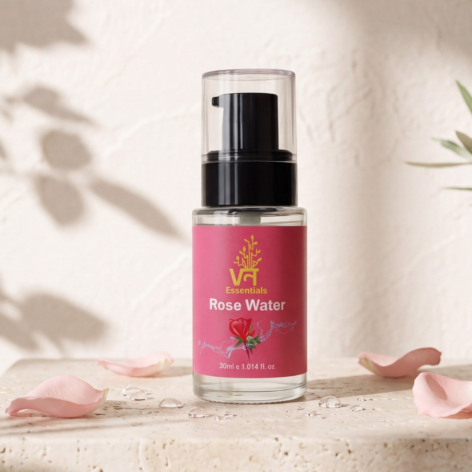 A 30ml bottle of Vann Essentials Rose Water with a pink label and black pump, set on a textured beige stone surface with scattered pink rose petals against a textured cream wall with soft leaf shadows.